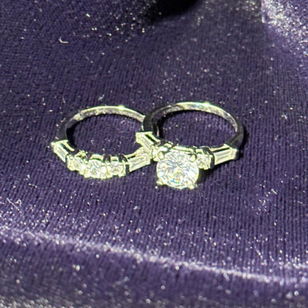 Two-piece sterling silver bridal set featuring a round brilliant-cut center stone with a shimmering halo setting, paired with a matching pavé-style eternity wedding band, displayed against a soft a purple textured surface