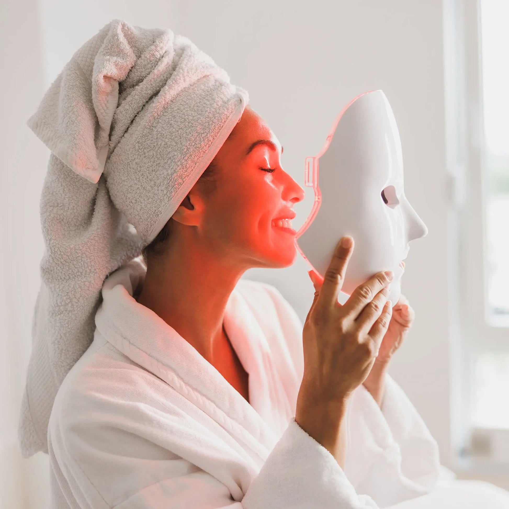 Woman in a bathrobe holding a white face mask to her face with a towel on her head.