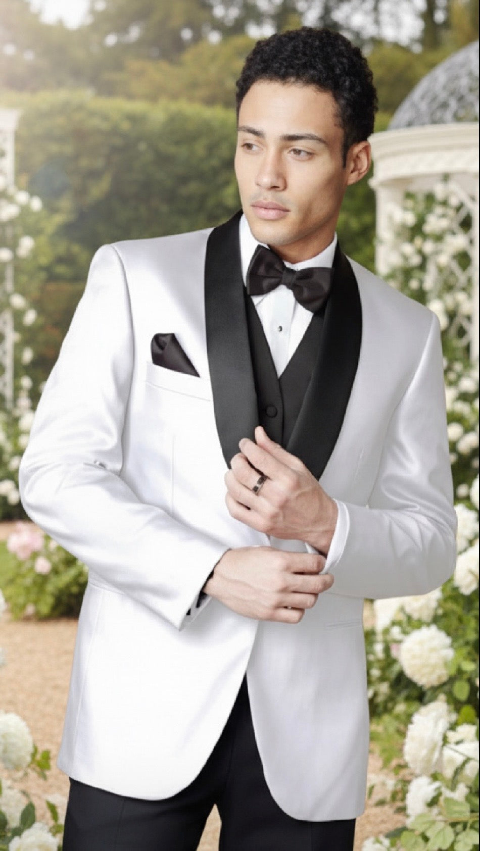 Man in a white tuxedo with black lapels and bow tie standing outdoors with greenery and flowers in the background.