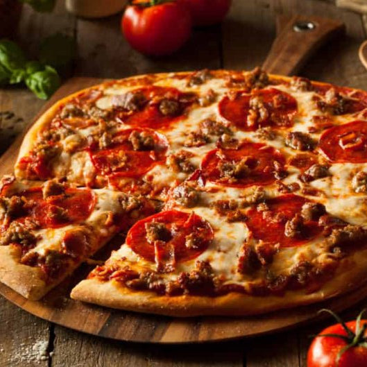 Personal-sized pizza with pepperoni and sausage on a wooden board with tomatoes and basil in the background.