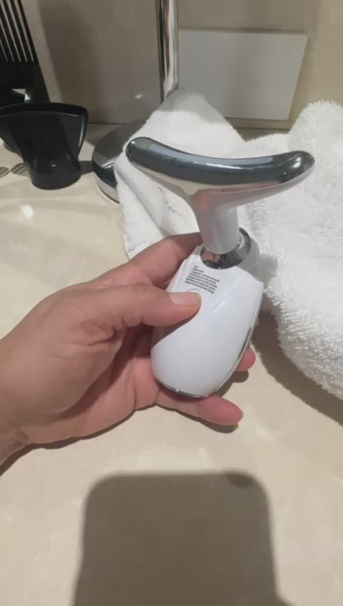 Handheld LED neck and face lifting therapy device for skin tightening and anti-aging massage. on a bathroom counter with a towel and sink in the background.