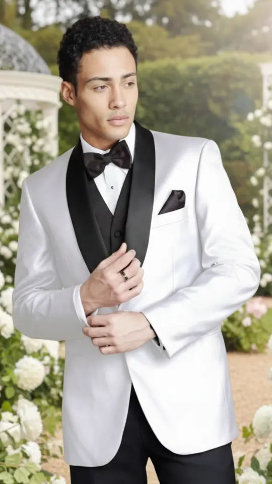 Man in a white tuxedo with black lapels standing outdoors with greenery and flowers in the background